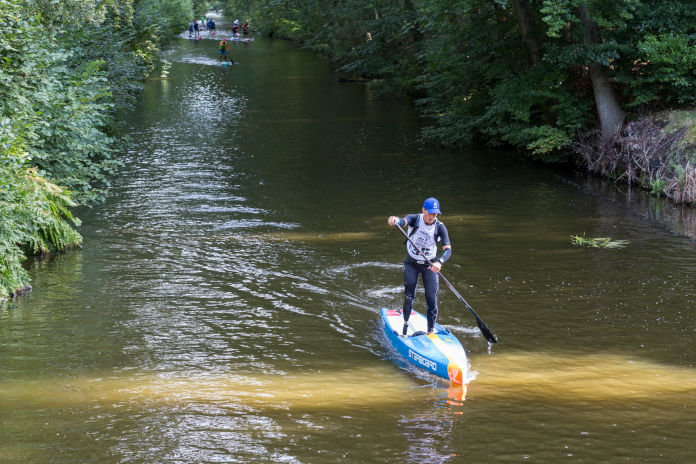 Eine Frau mit Beinprothese paddelt stehend auf einem Board, auf einem Fluss. Links und rechts Grünes. 