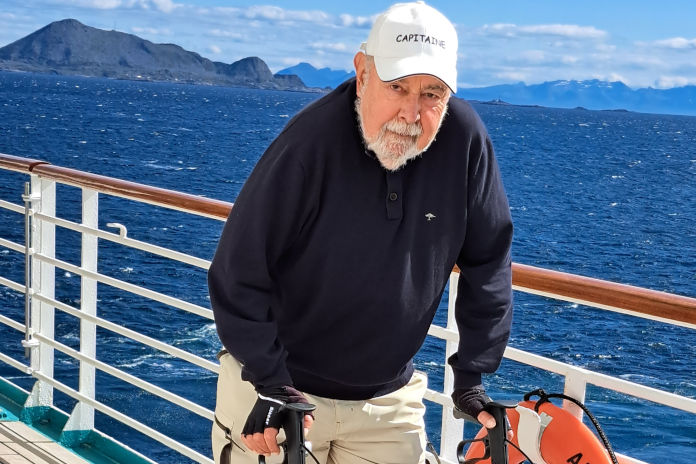 Ein Portraitfoto. Ein älterer Mann auf einem Kreuzfahrschiff. Er trägt eine Cappy. Im Hintergrund das Meer und Berge. Am Geländer hängt ein Rettungsring.