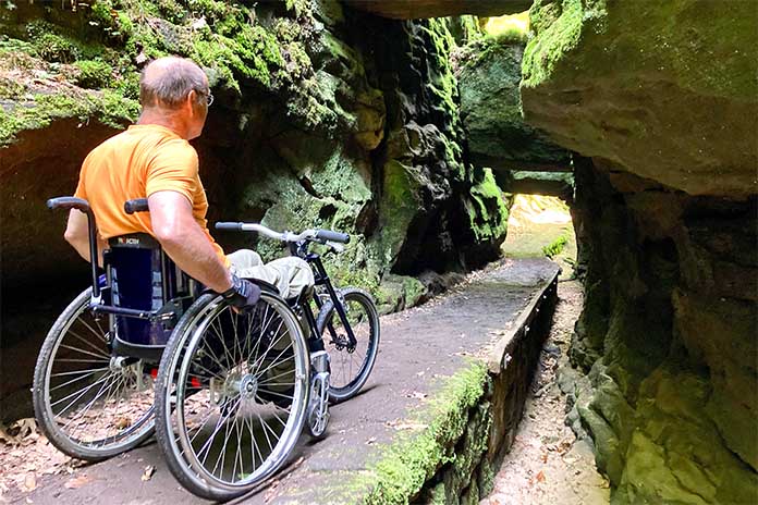 Ein Mann sitzend in einem Rollstuhl auf einem schmalen Felsenweg. Am Ende des Weges befindet sich ein Felsentor.