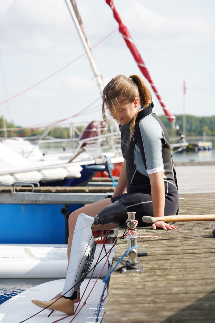 Ein junges Mädchen auf einem Holzsteg sitzend. Im Hintergrund befinden sich Segelboote. Sie trägt links eine Beinprothese.