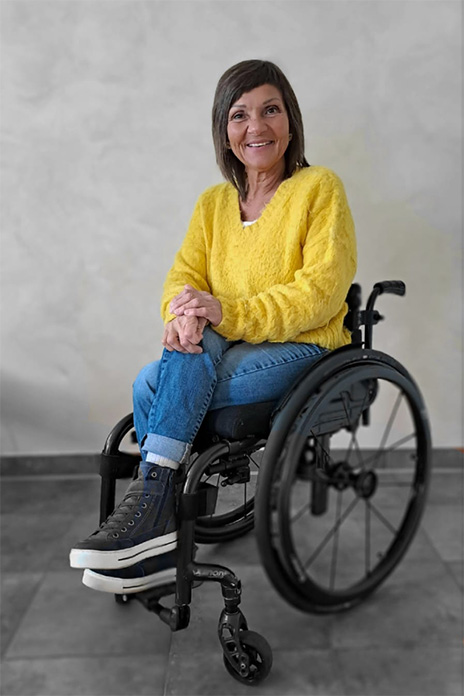 Eine Frau in einem schwarzen Rollstuhl sitzend. Sie lächelt, trägt einen gelben Pullover, Jeans und Boots. Die Beine sind übereinandergeschlagen. 