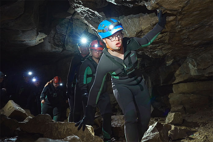 Mehrere Menschen in Neoprenanzügen, Helmen und Stirnlampen klettern durch eine Höhle.