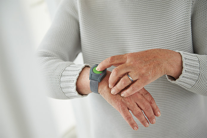 An einem rechten Handgelenk einer Frau ist ein Armband mit einem großen grünen Knopf. Mit dem Zeigefinger der linken Hand drückt sie den Notrufknopf.
