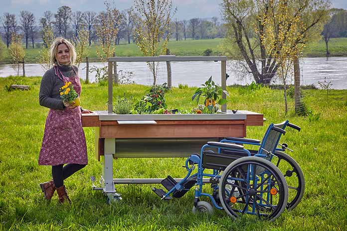 Ein mobiles, unterfahrbares und bepflanztes Hochbeet in der Natur. Vor dem Hochbeet steht ein Rollstuhl. An der linken Seite steht eine Frau mit einer Schürze. Sie hält in der rechten Hand eine Blume.