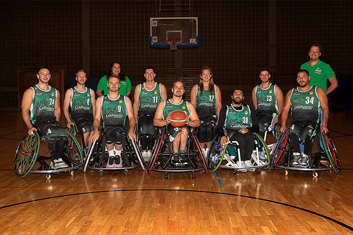 Ein Gruppenfoto. Mehrere Rollstuhlbasketballfahrer darunter auch eine Frau in zwei Reihen aufgeteilt. Ein Mann vorne in der Mitte sitzend hält einen Basketball in den Händen.