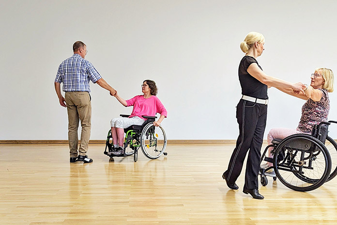 Zwei Rollstuhlfahrerinnen jeweils in einem Rollstuhl sitzend. Sie führen das Rollstuhltanzen aus. Die Dame rechts tanzt mit einer Frau. Die andere Frau im Rollstuhl tanzt mit einem Mann.