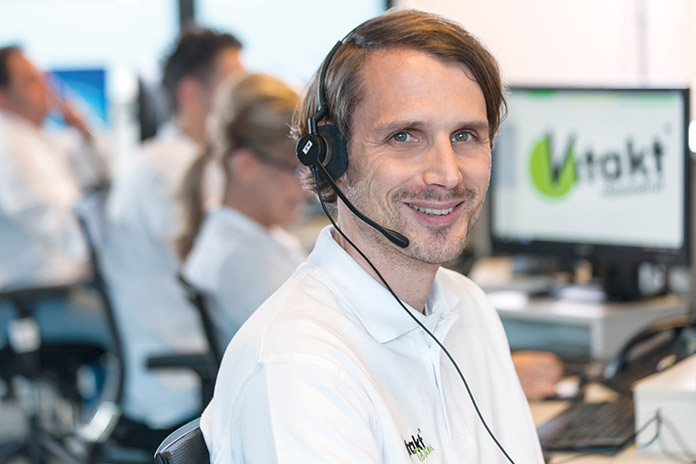 Ein männlicher Mitarbeiter mit weißem Poloshirt der Firma Vitakt. Er trägt ein Headset und lächelt in die Kamera.