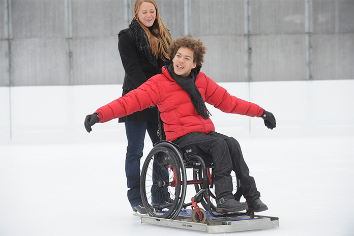 Auf einer Eisbahn. Ein sogenannter Eisgleiter, ein Untergestell für einen Rollstuhl um sich auf einer Eisbahn fortzubewegen. Auf dem Eisgleiter steht ein Rollstuhl in dem sitzt ein junger Mann. Er streckt beide Arme aus und wird von einer jungen Frau angeschoben.