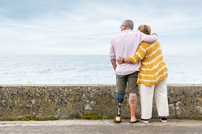 Vor einer Steinmauer steht ein Ehepaar und schaut aufs Meer. Der Mann ist am linken Bein amputiert und trägt eine Prothese. Er legt seinen rechten Arm um seine Frau. Sie legt ihren linken Arm auf seinen Rücken.