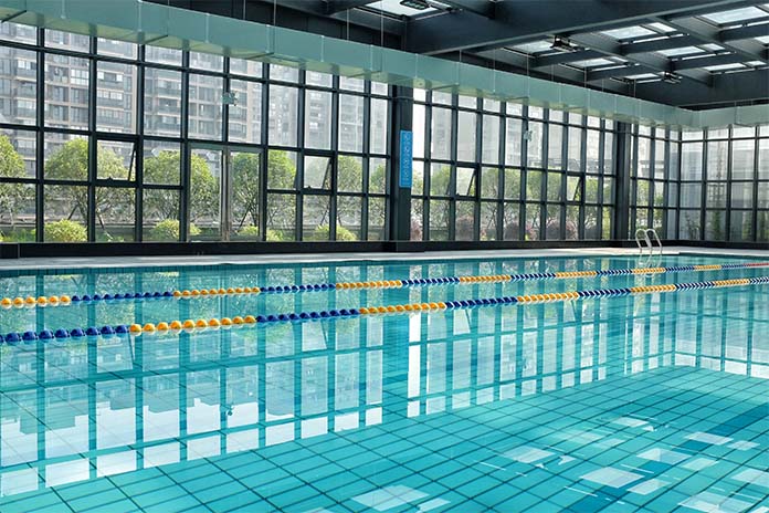 Ein Schwimmbad. Ein Teil des Schwimmbeckens ist als Bahn abgeteilt. Am Gebäude nach außen ist eine große Fensterfront mit abgeteilten Fenstern. 
