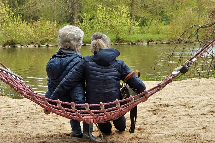 Zwei Frauen sitzen an einem Ufer eines Flusses in einer Hängematte aufrecht neben einander. 