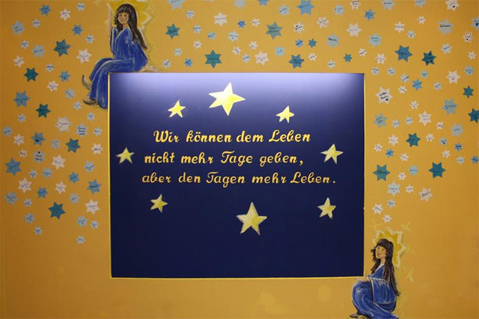 Auf einem dunkelblauen Hintergrund steht in gelben Buchstaben ein Spruch umgeben von Sternen. Oben links und unten rechts befinden sich jeweils ein Engel. Dahinter ein gelber Hintergrund mit lauter Sternen.