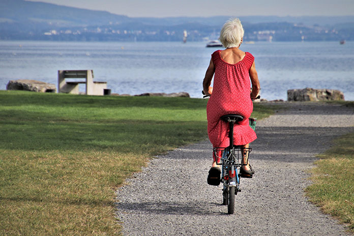 Das Bild zeigt eine ältere Frau, die in einem Park Fahrrad fährt. Sie trägt ein rotes Kleid und fährt auf einem Fahrrad mit einem Korb auf der Rückseite. Der Parkweg führt entlang eines Grünstreifens mit Gras und Bäumen. Im Hintergrund ist ein See oder Meer zu sehen, sowie einige Boote und eine Uferpromenade. Die Frau genießt offensichtlich die Fahrt und die entspannende Umgebung des Parks.