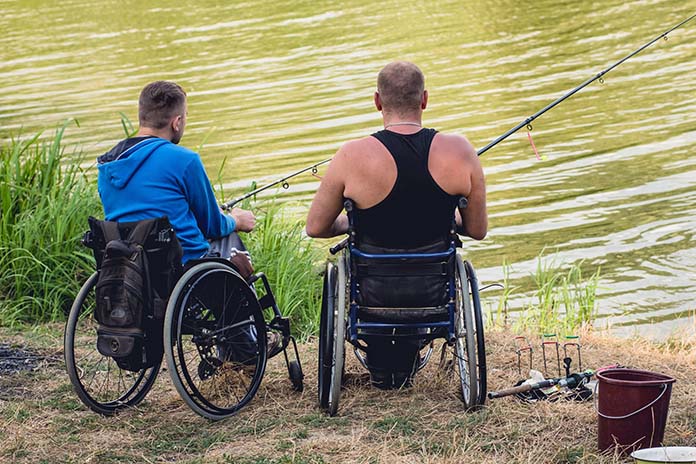 An einem Gewässer. Zwei Männer im Rollstuhl. Sie angeln. Auf dem Gras liegen weitere Angeln und zusätzlich steht dort ein Eimer.