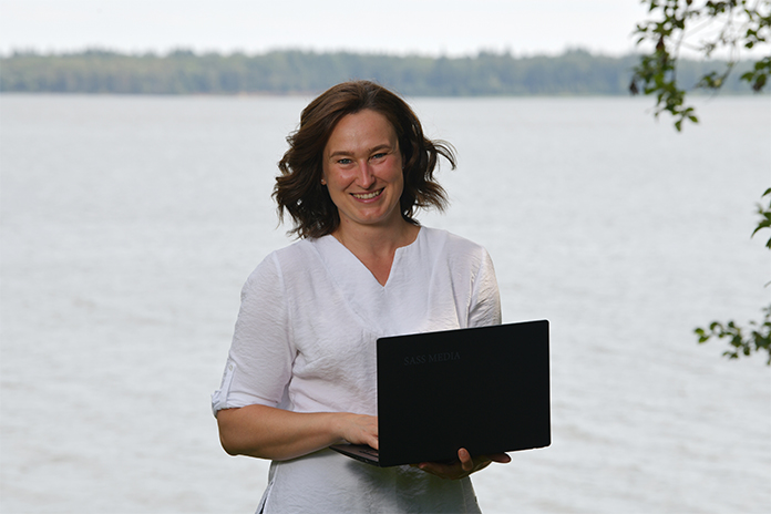 Portraitfoto von Lydia Saß, sie trägt eine weiße Bluse und vor sich einen aufgeklappten Laptop. Im Hintergund ist Wasser. 