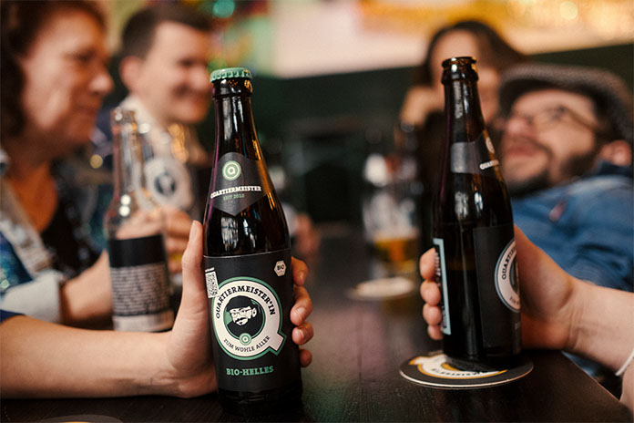 Zwei Bierflaschen werden von zwei Händen gehalten, im Hintergrund Menschen. 
