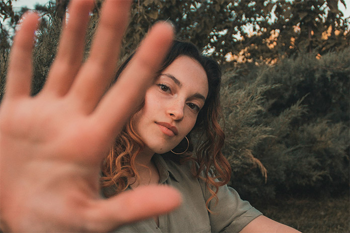 Eine junge Frau mit gelocktem Haar umgeben von Bäumen und Sträuchern. Sie hält ihre rechte Hand etwa auf Kopfhöhe. Die Finger hält sie zu einer fünf, leicht gespreizt. Ihren Kopf hält sie etwas seitlich.