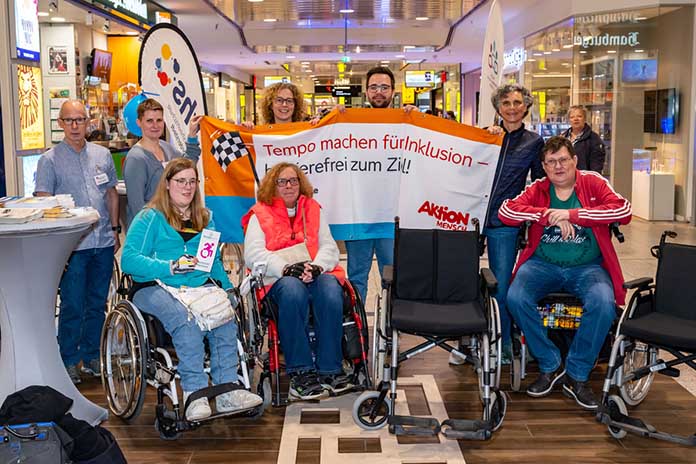 Zwei Reihen in der vorderen Reihe drei Personen in Rollstühlen. In der hinteren fünf Personen stehend mit einem Banner in der Hand. Auf der linken Seite steht ein Cocktailtisch mit Werbematerial.