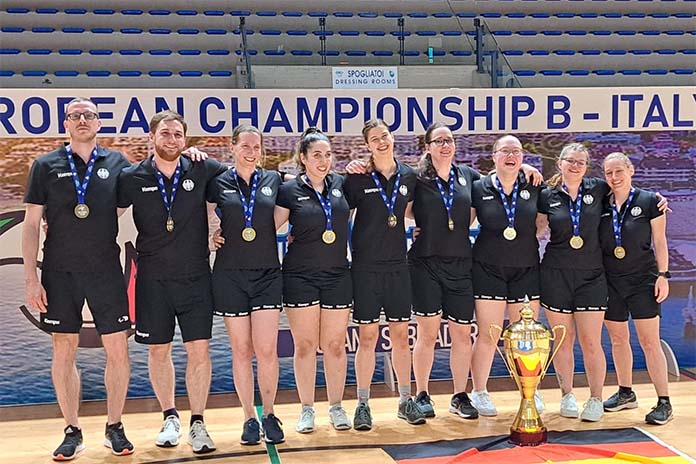 Ein Mannschaftsfoto eines Sportteams in einer Reihe. Sieben Frauen nebeneinander, vorne links stehen zwei Männer. Alle tragen kurze schwarze T-shirts und kurze schwarze Hosen. Im Vordergrund steht ein Pokal, alle tragen eine Medaille.