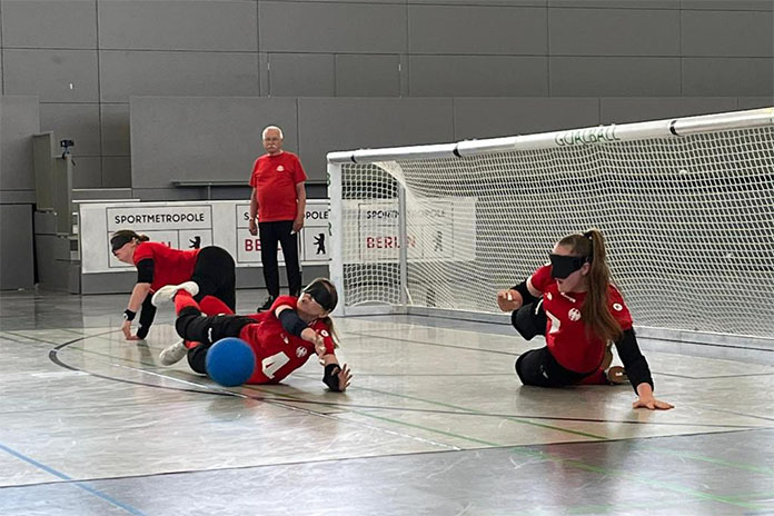 In einem Spiel in einer Sporthalle. Drei Frauen vor einem Tor, zwei von ihnen liegend. Die Frau in der Mitte (liegend) versucht einen blauen Ball zu fangen. Alle tragen eine schwarze Maske vor den Augen. Links außen am Tor steht ein Mann.