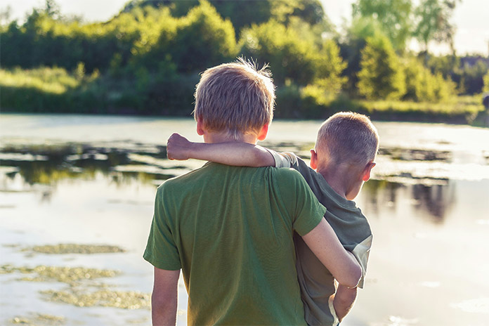 Zwei Jungs an einem Gewässer. Der größere Junge hält den kleinen Jungen im Arm. Der kleine Junge hält den größeren Jungen mit seinem linken Arm um den Hals fest.