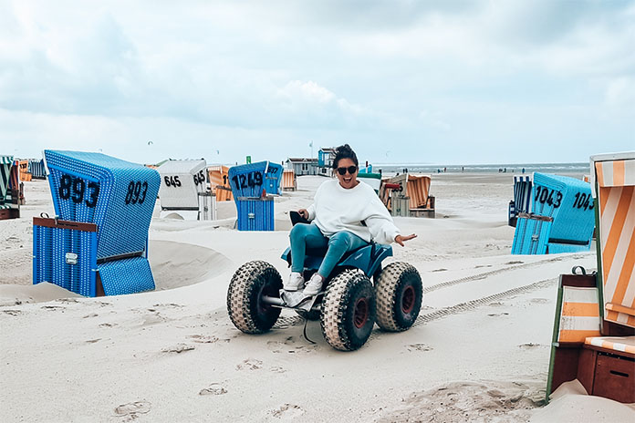 Ein Elektrostrandrolli - ein Fahrzeug mit Sitzfläche und vier großen Reifen am Strand zwischen Strandkörben fahrend. Das Fahrzeug wird von einer Frau gefahren, sie trägt eine Sonnenbrille und lacht. 