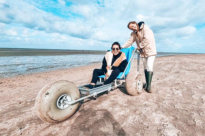 An einem Strand in einem Wattmobil bestehend aus Sitzfläche, einem großen Gummireifen vorne und zwei breiten Gummireifen hinten, sitzt eine Frau mit Sonnenbrille und lacht. Neben ihr steht eine weitere Frau in Gummistiefeln und hält sich an dem Wattmobil fest. 