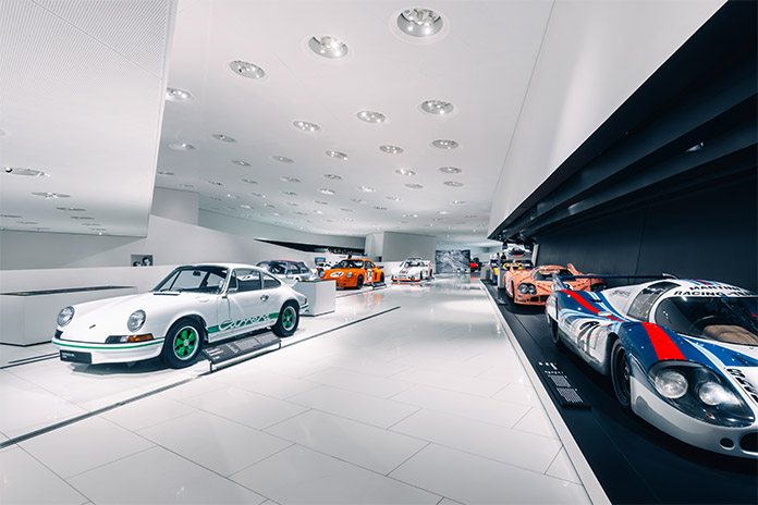Im Porsche Museum. Verschiedene Porsche Modelle unterschiedlich in ihrer Art und Form aufgestellt. Auf der rechten Seite befinden sich die Fahrzeuge hintereinanderstehend. Vor den Fahrzeugen befinden sich Informationsschilder.