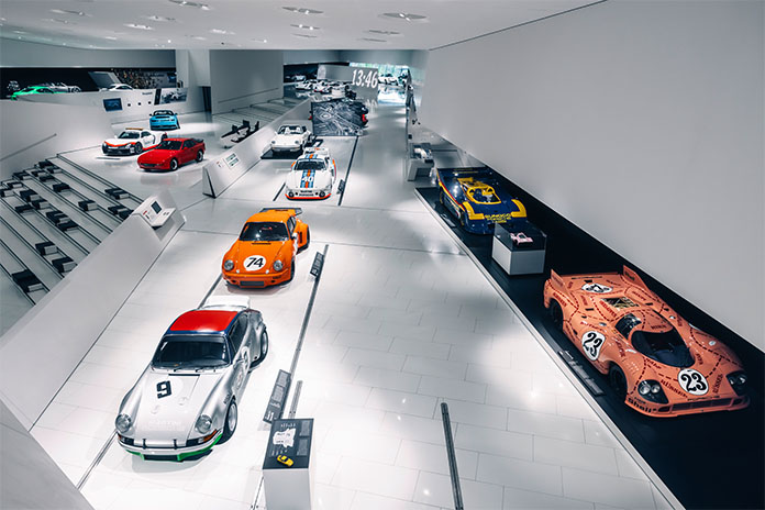 Im Porsche Museum. Verschiedene Porsche Fahrzeuge in verschiedenen Ausführungen wurden in das Gebäude integriert. Sie stehen teilweise hintereinander und nebeneinander. Vor den Fahrzeugen sind Informationsschilder zu den jeweiligen Autos.