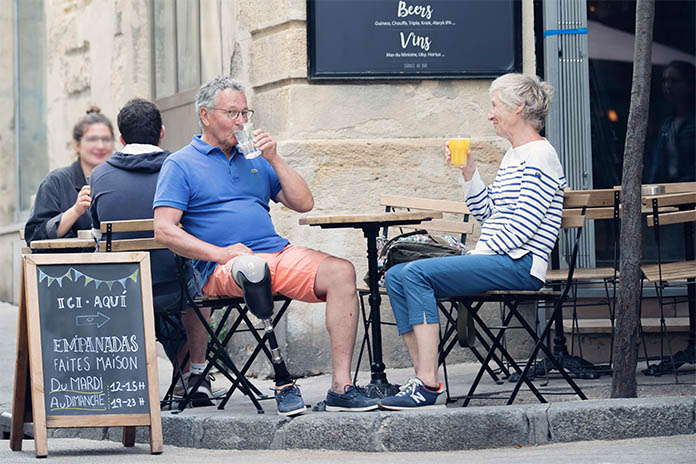 An einer Ecke eines Straßencafes sitzt ein Pärchen. Der Mann auf der linken Seite ist an seinem rechten Bein amputiert und trägt eine Prothese. Die Frau sitzt rechts neben ihm. Beide trinken etwas.