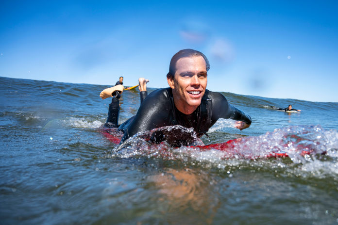 Ein Mann liegt auf einem Surfbrett auf dem Meer und paddelt. Er trägt links eine Fußprothese. Hinter ihm ist eine Welle und zwei andere Surfer zu sehen. 