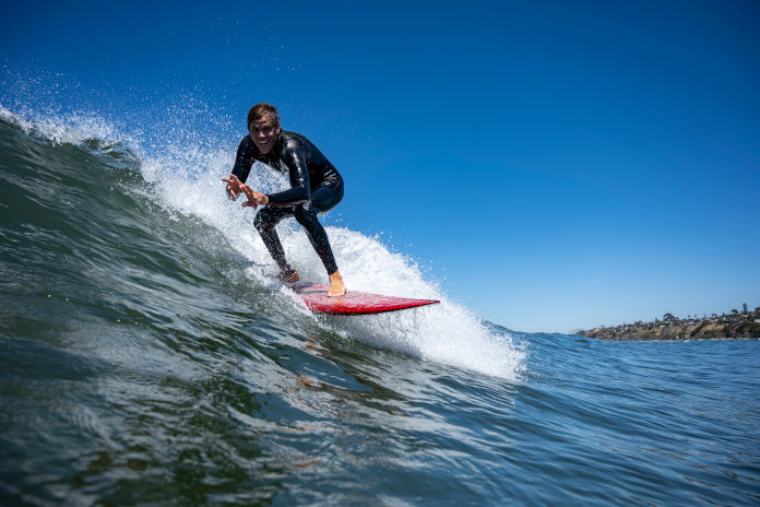 Ein junger Mann leicht gebeugt, stehend auf einem roten Surfbrett. Er lacht und reitet auf einer Welle.