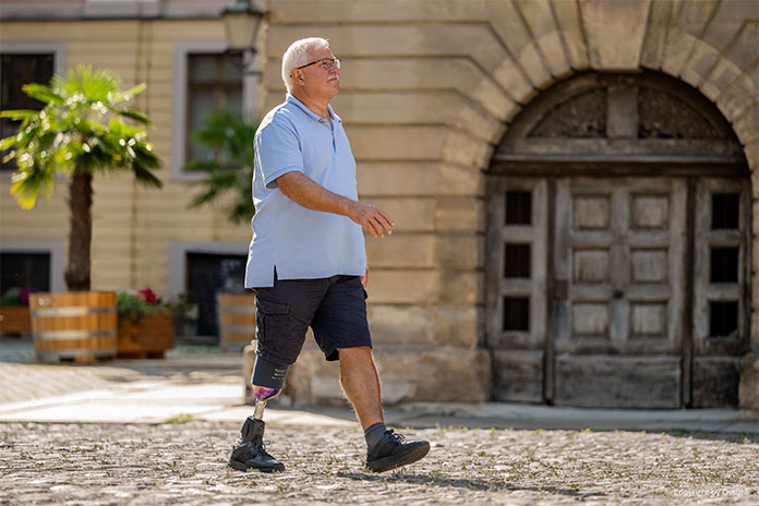 Ein älterer Mann läuft durch eine Altstadt. Sein rechter Arm schwingt dabei nach vorne. Die Sonne scheint. Er trägt am rechten Bein eine Prothese. 