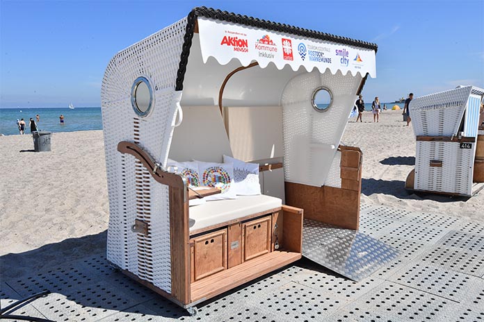 Ein seitlich aufgenommenes Foto von einem rollstuhlgerechten Strandkorb. Dieser steht auf einer befahrbaren Fläche an einem Strand. Auf der rechten Seite befindet sich eine Rampe und auf der linken eine Sitzfläche. Im Hintergrund ist das Meer. 