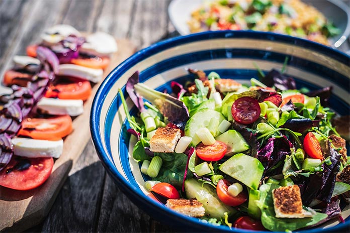  Eine bunte Salatschüssel mit Tomaten, Gurken und Blattsalaten, daneben Tomaten, Mozzarella und Zwiebeln auf einem Holzbrett.