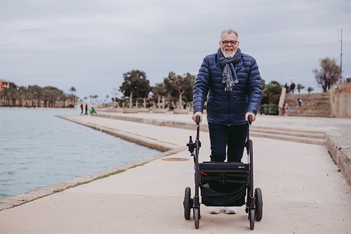 Ein älterer Mann mit seinem Rollator, stehend auf hellbeigefarbenen Beton. Rechts neben ihm befindet sich das Meer. Der Mann trägt eine Brille, eine dunkelblaue Jacke und einen Schal.