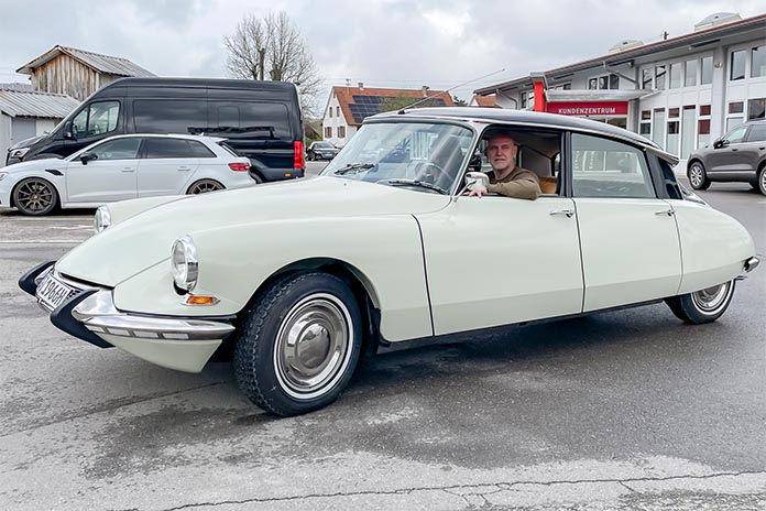 Ein Mann sitzt in einem cremefarbenen Citroën DS, der auf einer Straße geparkt ist. Im Hintergrund sind ein schwarzer Lieferwagen, ein weißes Auto und ein Gebäude mit der Aufschrift "KUNDENZENTRUM" zu sehen.