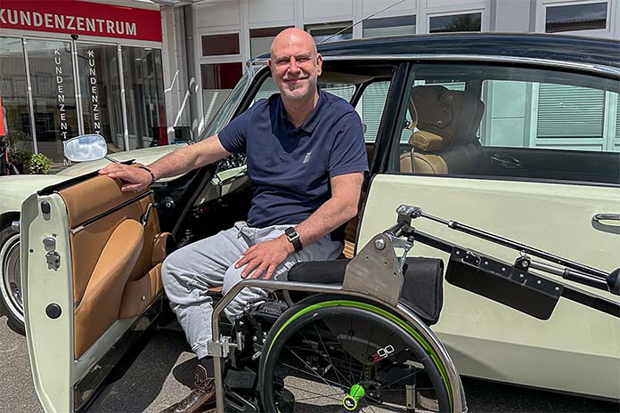 Ein Mann in einem CitroenDS sitzend auf einem Rutschbrett, er hält sich an der Tür fest. Vor ihm sein Rollstuhl. 