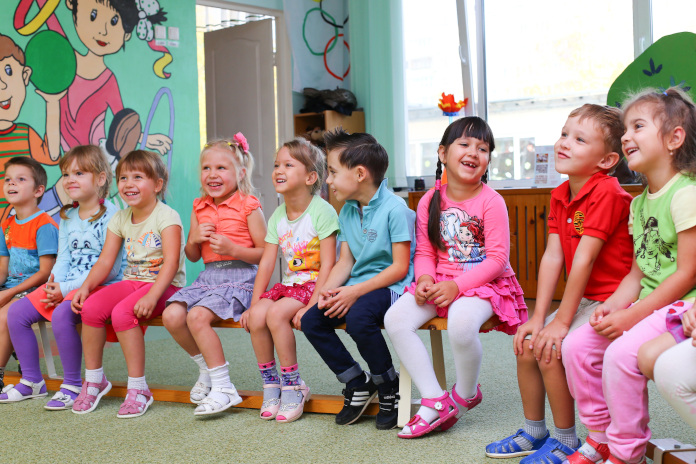 In einem Kindergarten. Mehrere Kinder sitzen nebeneinander auf einer Bank und lachen. 