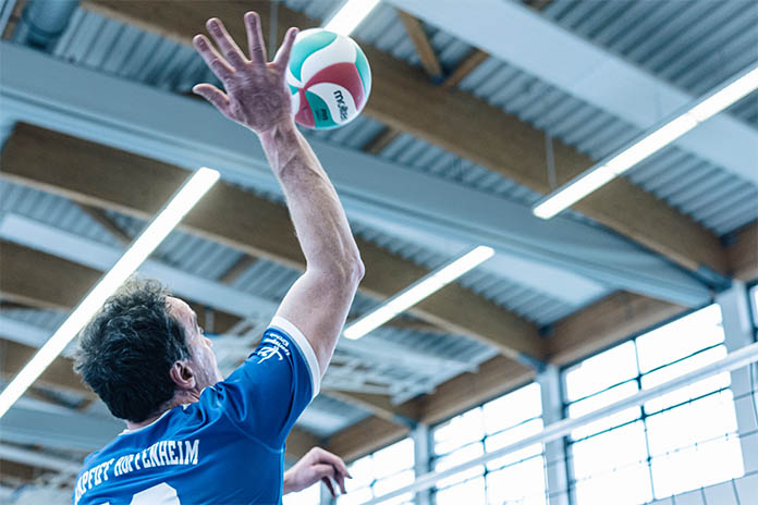 Ein Mann sitzt mit ausgestrecktem rechten Arm. In der Luft vor ihm ist ein Volleyball, bereit für einen Aufschlag, davor ist ein Volleyballnetz.