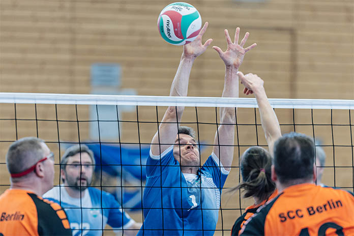 Mehrere Spieler spielen Sitzvolleyball. Zwei Teams spielen gegeneinander, dazwischen ist ein Volleyballnetz. Ein Spieler streckt beide Arme und Hände aus. Davor ist ein Ball. 