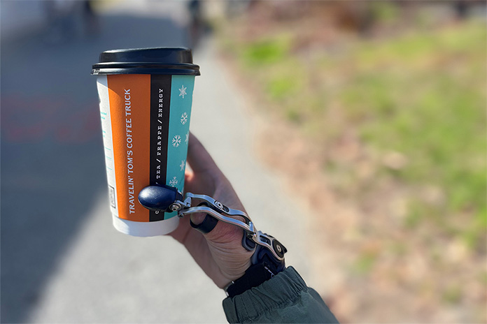 Eine Frauenhand mit Daumenprothese hält einen bunten Papp-Becher mit Deckel. 