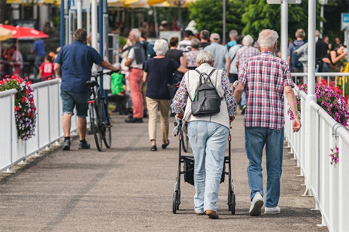 Auf einem Gehweg mit weißem Geländer spazieren zwei ältere Menschen. Die Fau auf der linken Seite mit einem Rollator und Rucksack. Rechts neben ihr ein Mann. Er hält sich mit seiner linken Hand am Rollator fest. Vor ihnen laufen mehrere Menschen.